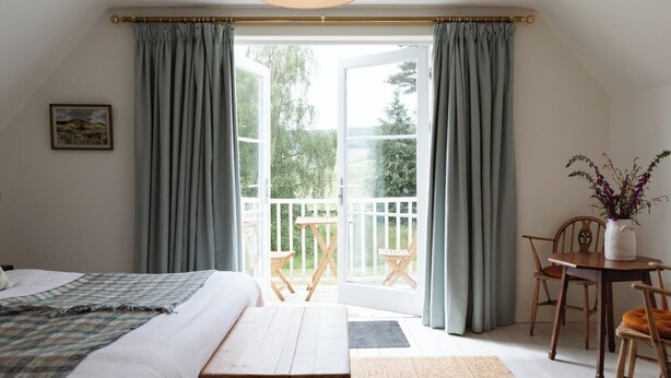 A bedroom with a bed, a wooden bench, a small table with a vase of flowers, and a balcony with a table and chairs. - Stunning 3 bedroom Cottage on a private estate in SW Scotland (Mossdale)