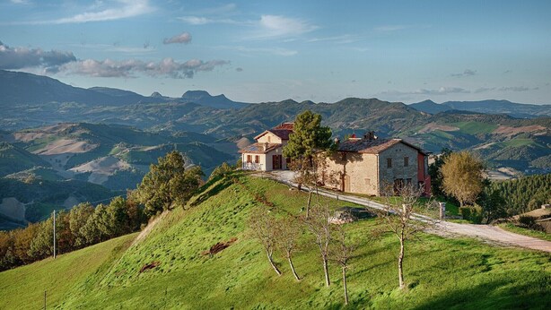 Une maison de campagne aux murs en pierre, entourée de collines verdoyantes et sous un ciel d’un bleu limpide.