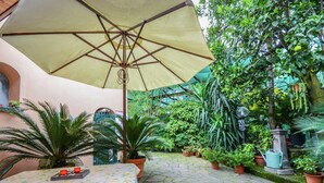 Une cour dallée de pierres, avec une table et des chaises placées sous un grand parasol, le tout entouré d’une végétation luxuriante et de plantes en pot.