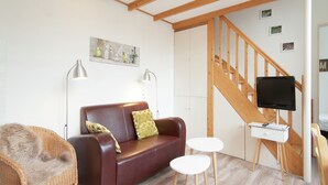 A cozy living room with a brown leather sofa, a wicker chair, a small white table, and a TV mounted on a wooden wall. - Apartment in Bergen aan Zee near the Beach (Bergen aan zee)