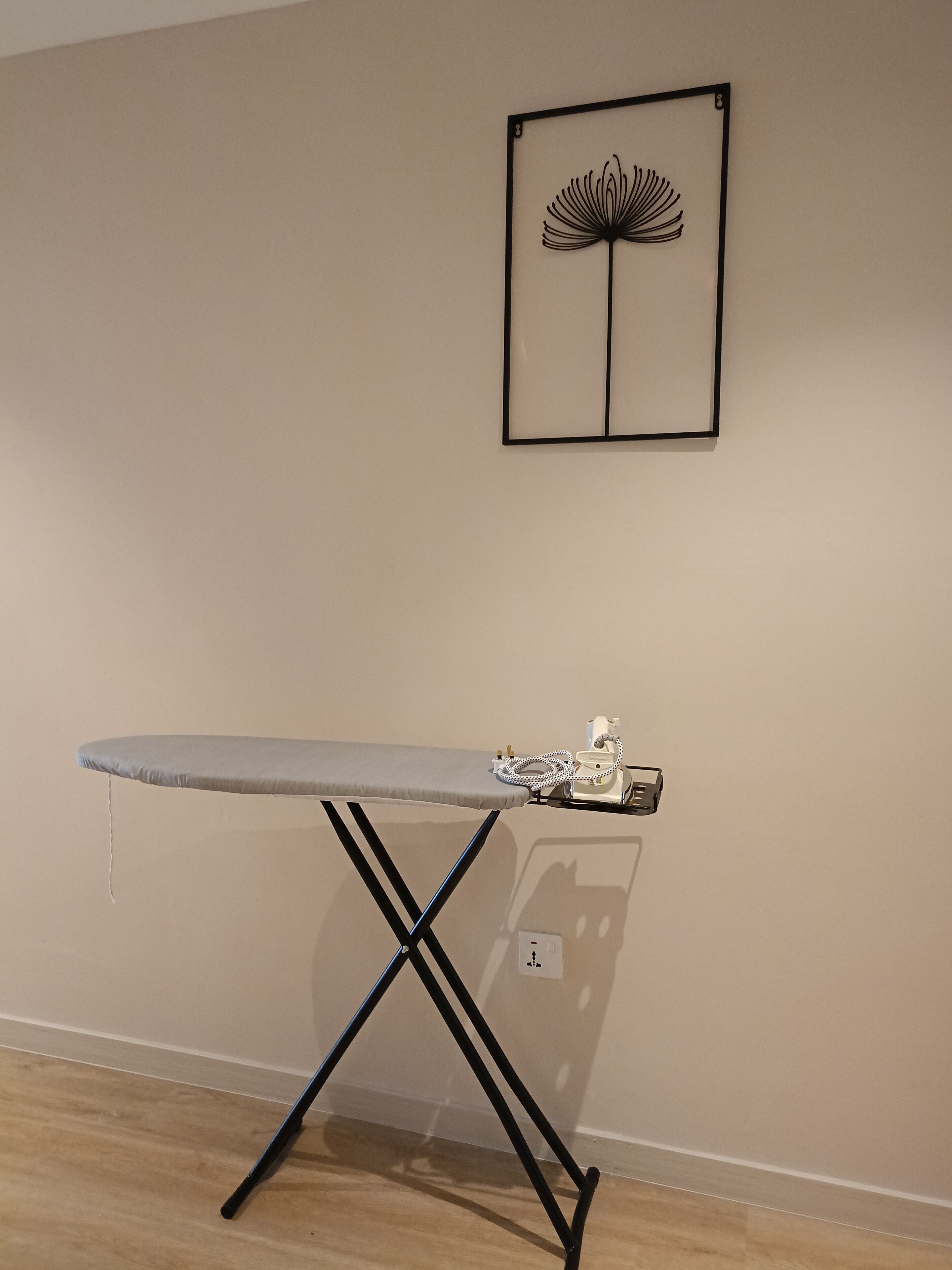 A room with a wooden floor, a grey ironing board with a white corded iron, and a framed artwork on the wall.