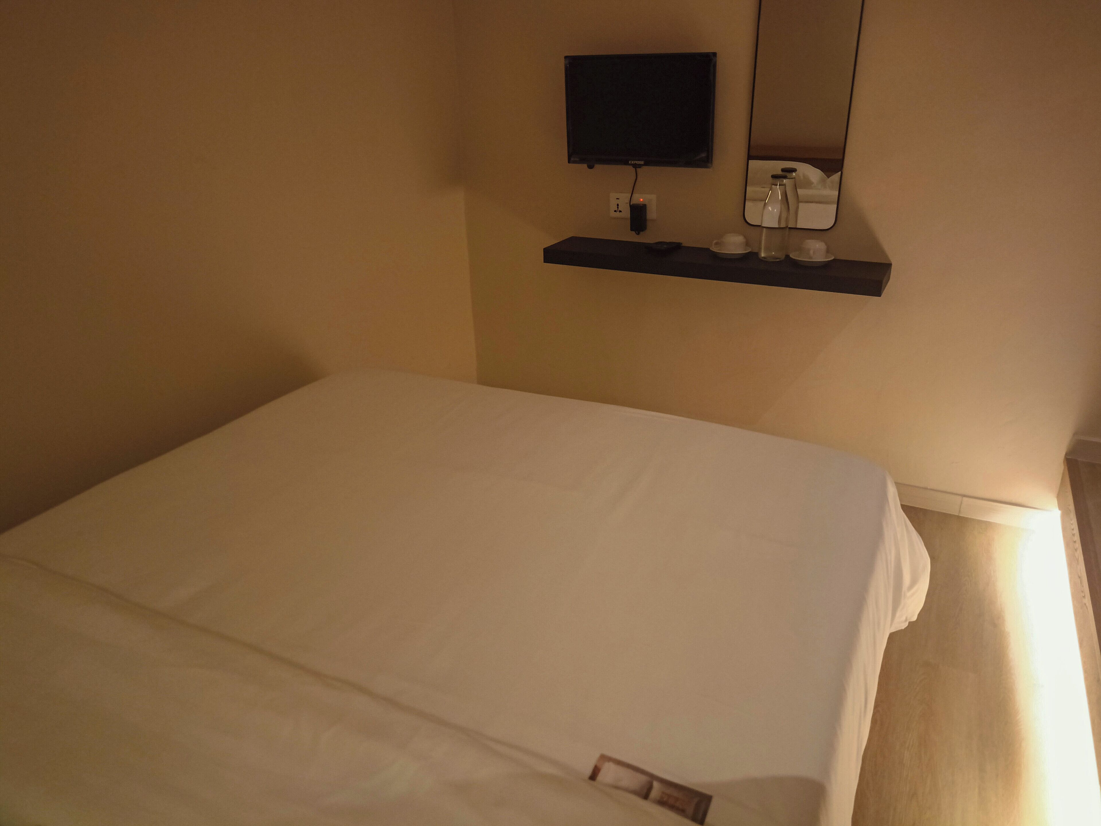 Una cama bien hecha en una habitación de hotel, con un televisor montado y una pequeña estantería con un control remoto, tazas y una botella.