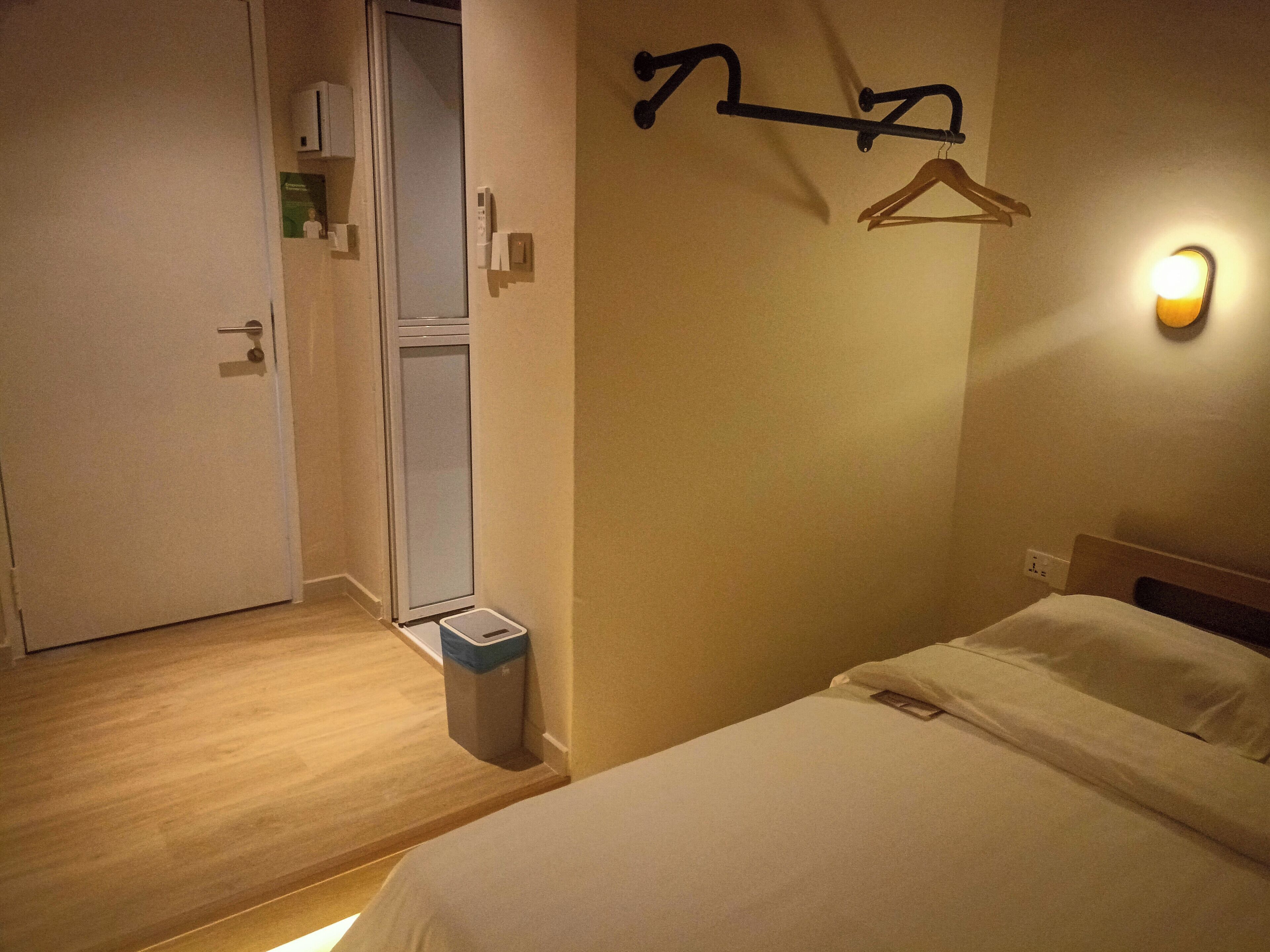 A hotel room with a bed, a wardrobe, a trash bin, and a wall-mounted light fixture.