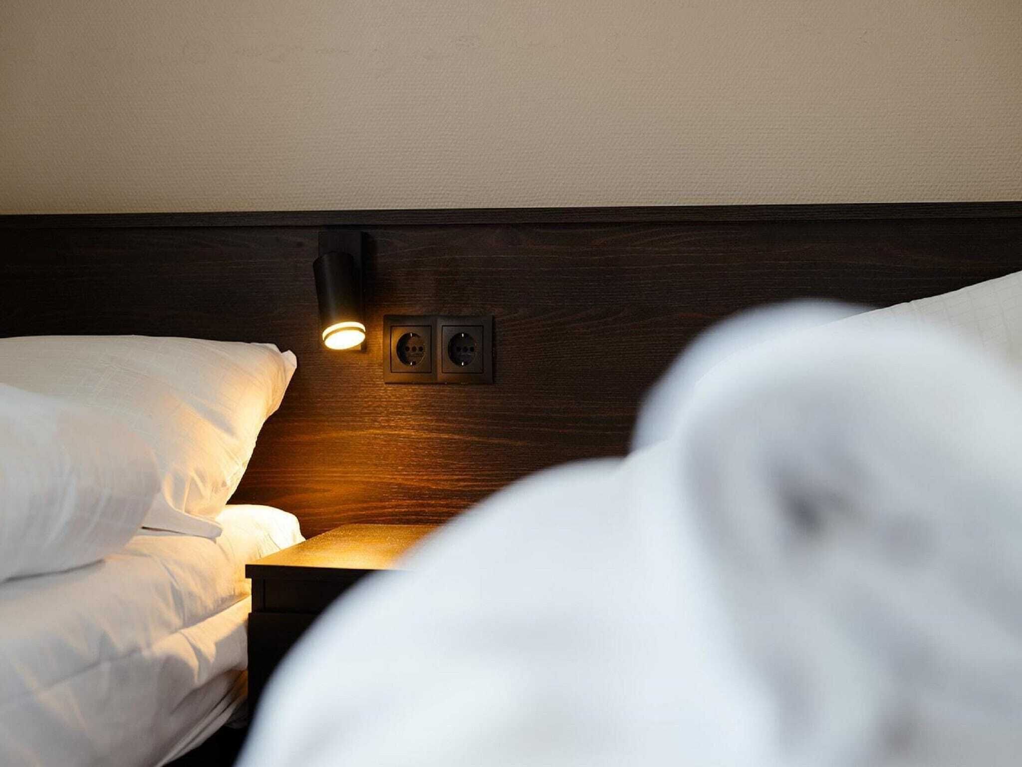 Een hotelkamer met een houten hoofdeinde, een nachtkastje met een lamp en een bed met wit beddengoed.