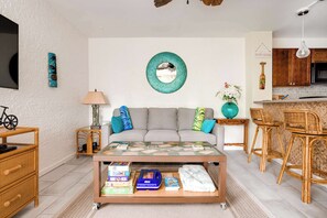 Una sala de estar con un sofá, una mesa de centro y sillas de mimbre. Hay una zona de cocina con armarios de madera y una lámpara colgante.