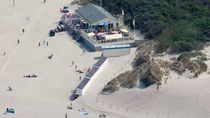 Ein Luftbild eines an der Küste stehenden Gebäudes mit einer langen Holzterrasse, die sich auf den Strand erstreckt, umgeben von Badegästen und ausgestattet mit Sonnenschirmen und Sitzbereichen.