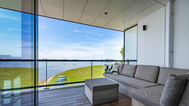 A modern living room with a large glass window overlooking a body of water, a wooden deck, and a beige sofa. - Apartment in Veere with Lake Views & Marina (Kamperland)