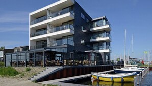 A modern multi-story building with balconies, a covered outdoor seating area, and a dock with boats. - Apartment in Veere with Lake Views & Marina (Kamperland)
