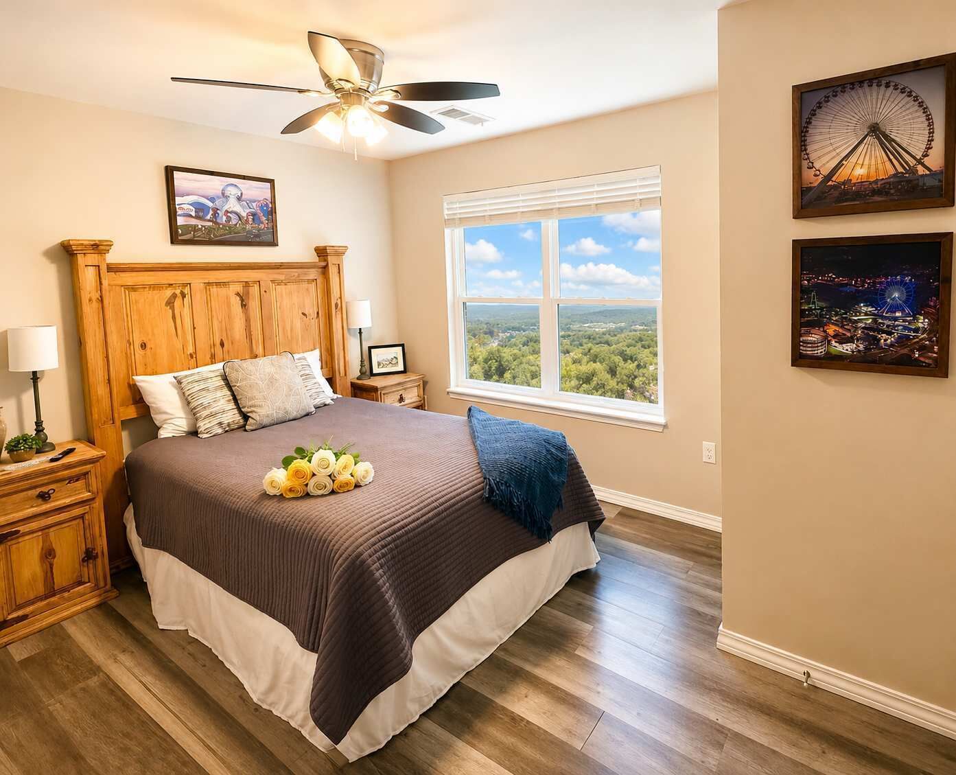 Une chambre à coucher avec une tête de lit en bois, une grande fenêtre donnant sur un beau paysage, un ventilateur de plafond et des tableaux encadrés accrochés au mur.