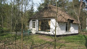 Een klein wit huis met rieten dak, omringd door bomen en een gazon.