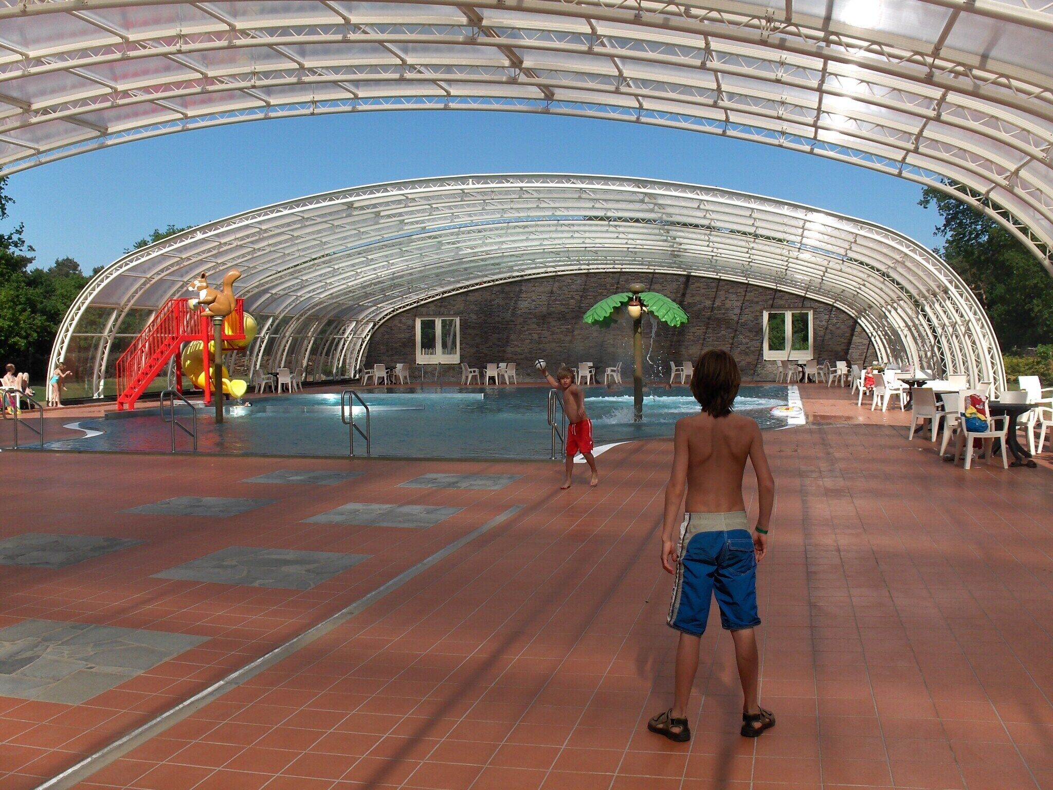An indoor swimming pool with a curved roof structure, surrounded by seating areas and a water slide.