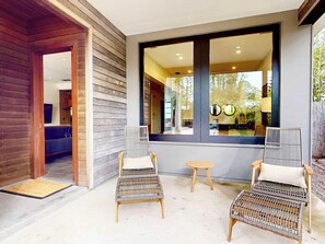 Un espace extérieur moderne aménagé avec des chaises en osier et une petite table, une grande fenêtre donnant sur l’intérieur et une porte en bois menant à l’intérieur.