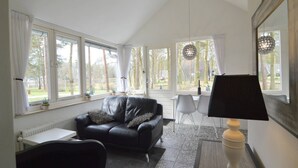 Una sala de estar moderna con un sofá de cuero negro, una mesa de comedor con dos sillas y una lámpara. Amplias ventanas ofrecen vistas al exterior.