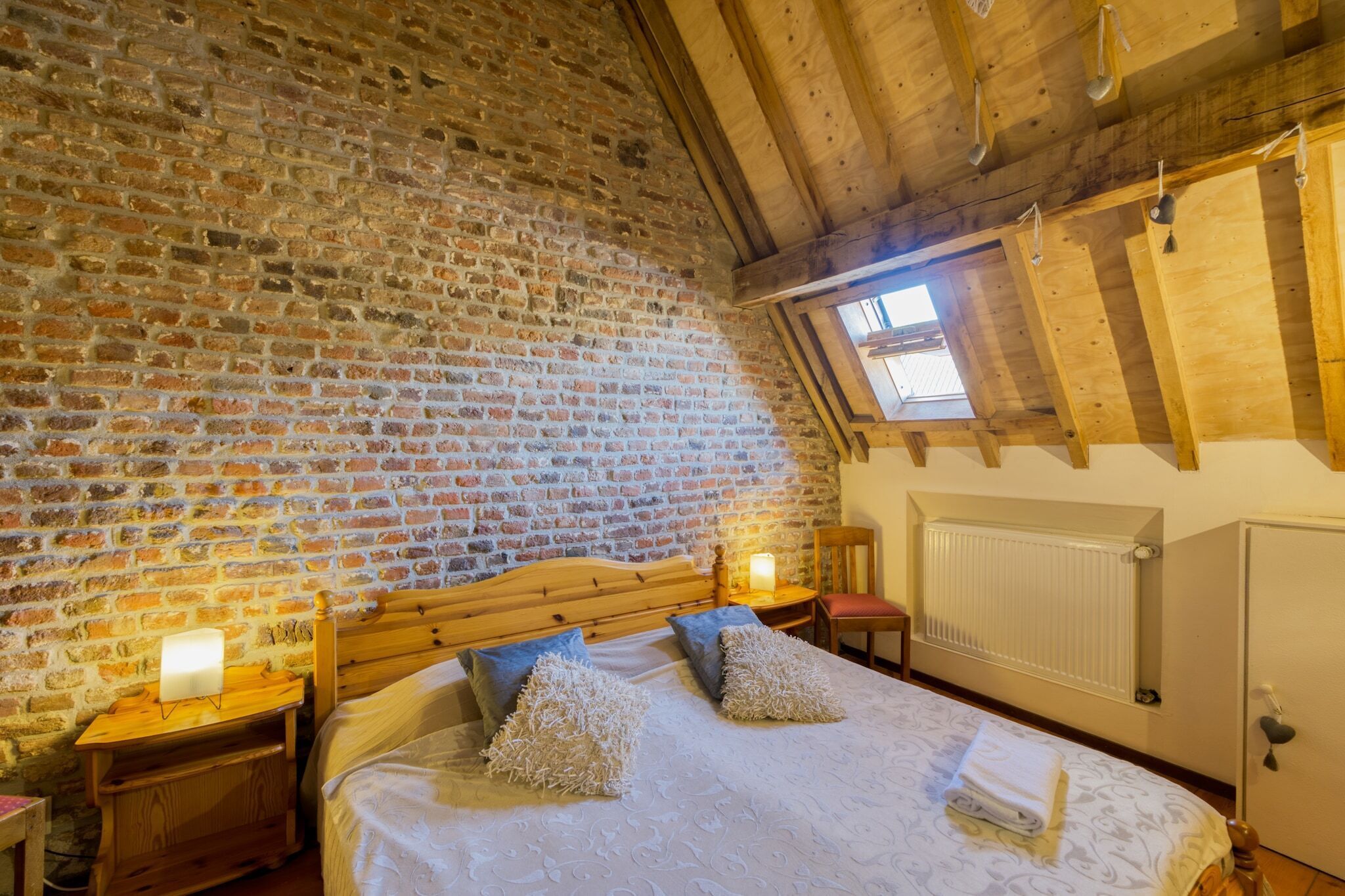 A bedroom with a brick wall, wooden ceiling beams, a bed with pillows, a nightstand with a lamp, and a window with blinds.