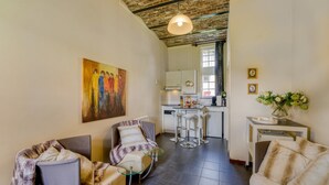 Ein modernes Wohnzimmer mit Sofa, Sessel und einem Glastisch. An der Wand hängt ein Gemälde, im Hintergrund ist eine Küchenzeile zu sehen.