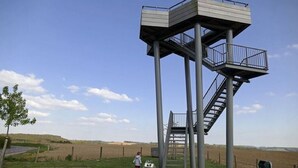 Ein metallener Aussichtsturm mit Treppe und Plattform, umgeben von offenen Feldern und blauem Himmel.