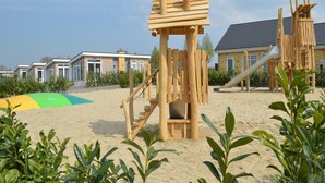A wooden play structure in a sandy area with a building in the background. - Charming Retreat for Your Escape (Bemelen)
