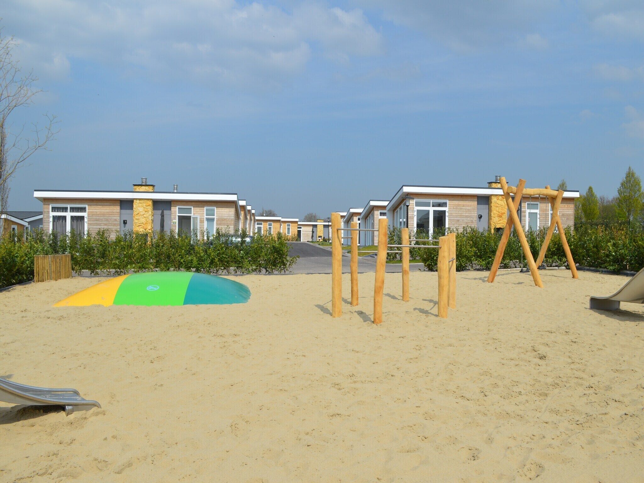 A sandy playground area with a swing set and a slide, surrounded by modern buildings with large windows and a stone facade.