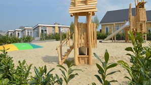 Un patio de juegos con estructuras de madera para escalar y un tobogán, rodeado de arena y follaje.