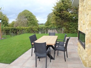 Un patio exterior con una mesa y sillas de madera, un muro de piedra y un seto al fondo.