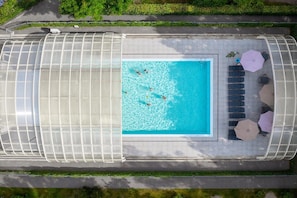 An aerial view of a swimming pool with a glass enclosure, surrounded by lounge chairs and umbrellas.