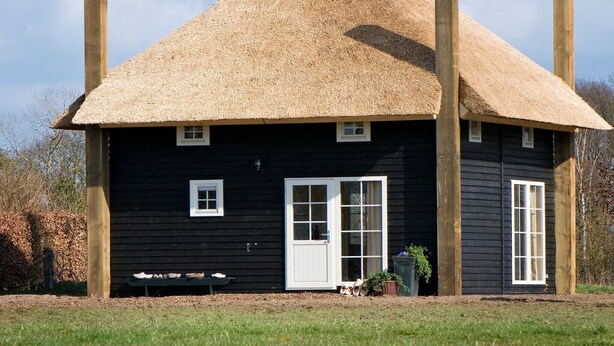 Una casa negra de fusta amb teulada de palla, finestres blanques i una porta.