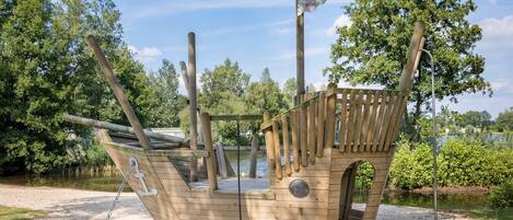 A wooden pirate ship play structure with a ladder, a steering wheel, and a flag.