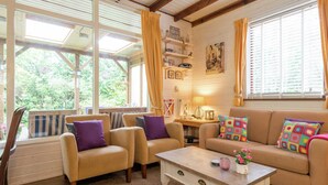 A cozy living room with a sofa, two armchairs, a coffee table, and a wooden side table. There are windows with curtains, a bookshelf, and a wall-mounted shelf with decorative items.