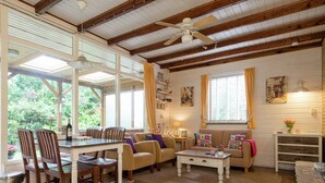 A spacious living area with a dining table, chairs, a sofa, and a coffee table. There is a ceiling fan, large windows with curtains, and a view of greenery outside.