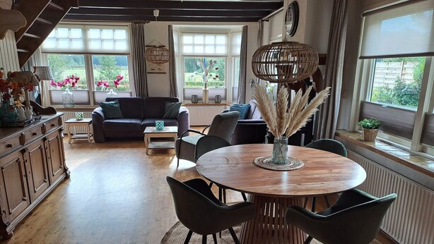 A spacious living room with a wooden dining table surrounded by chairs, a sofa, and a coffee table. Large windows provide natural light and views of greenery outside. - Holiday Home in Nijeberkoop near Nature (Nijeberkoop)