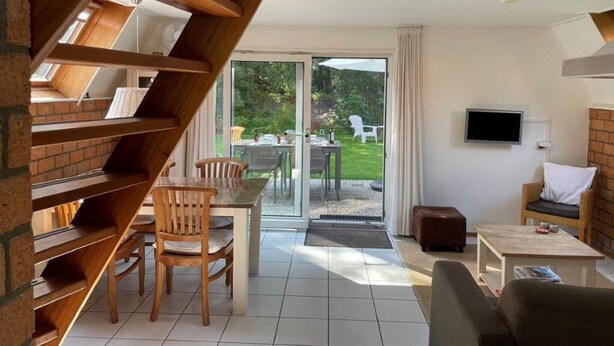 A modern living room with a staircase, a dining area, and a view of the garden through glass doors.