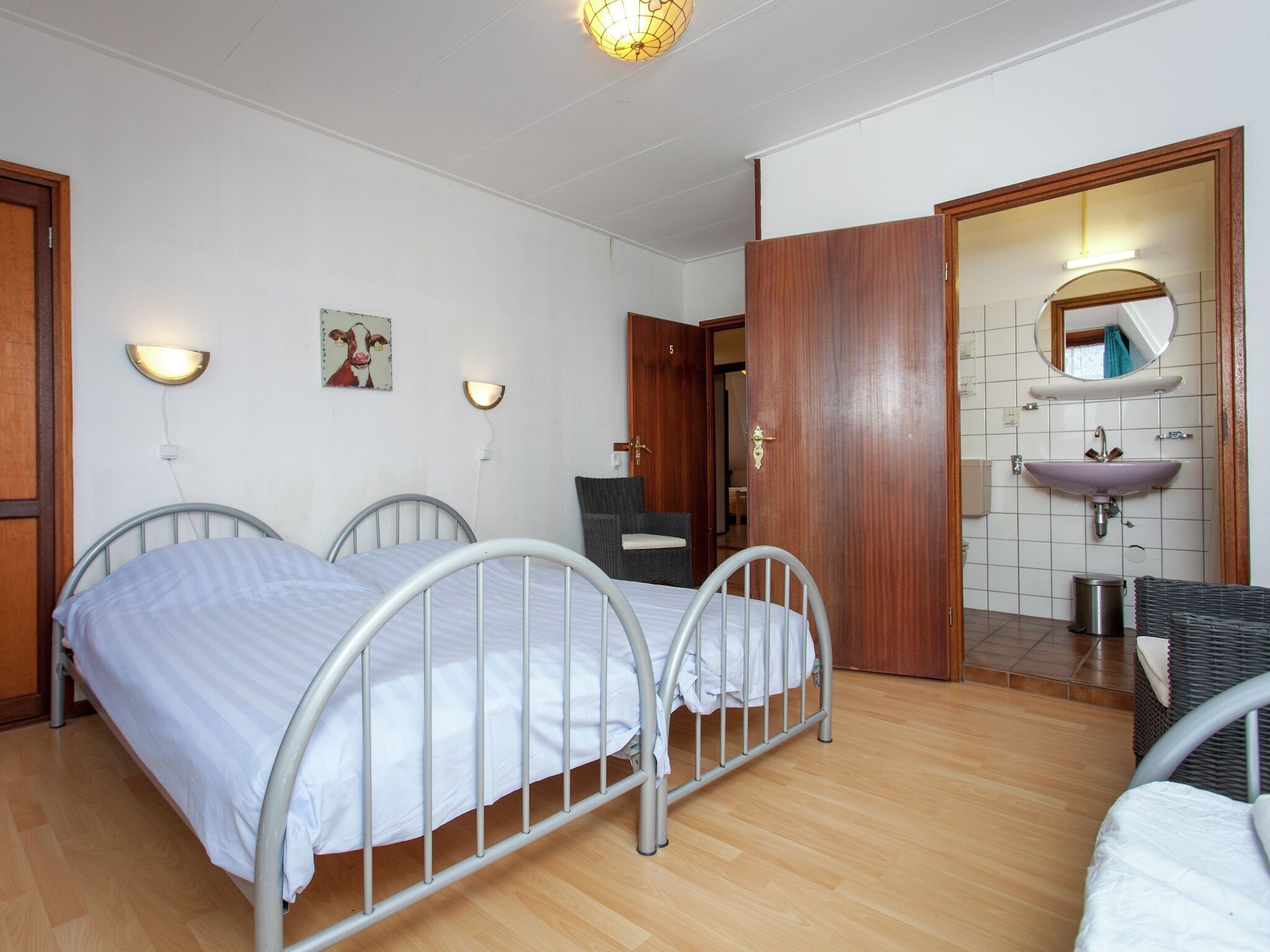 Ein Zimmer mit zwei Betten aus Metall, einer Holztür, einem Badezimmer mit Waschbecken und Spiegel sowie Wandleuchten.