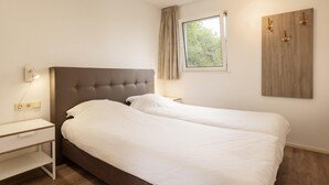 A bedroom with a double bed, a bedside table, a lamp, a window with curtains, and a wooden wall art piece.