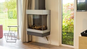 A corner fireplace with a glass enclosure, a small table, and a view of greenery outside.