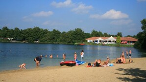 Una spiaggia sul lago con persone che nuotano e prendono il sole, una barca e un edificio con tetto rosso sullo sfondo.