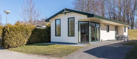 Un edificio de un piso, de color blanco, con techo verde y ventanas amplias, que cuenta con un patio con mobiliario exterior.