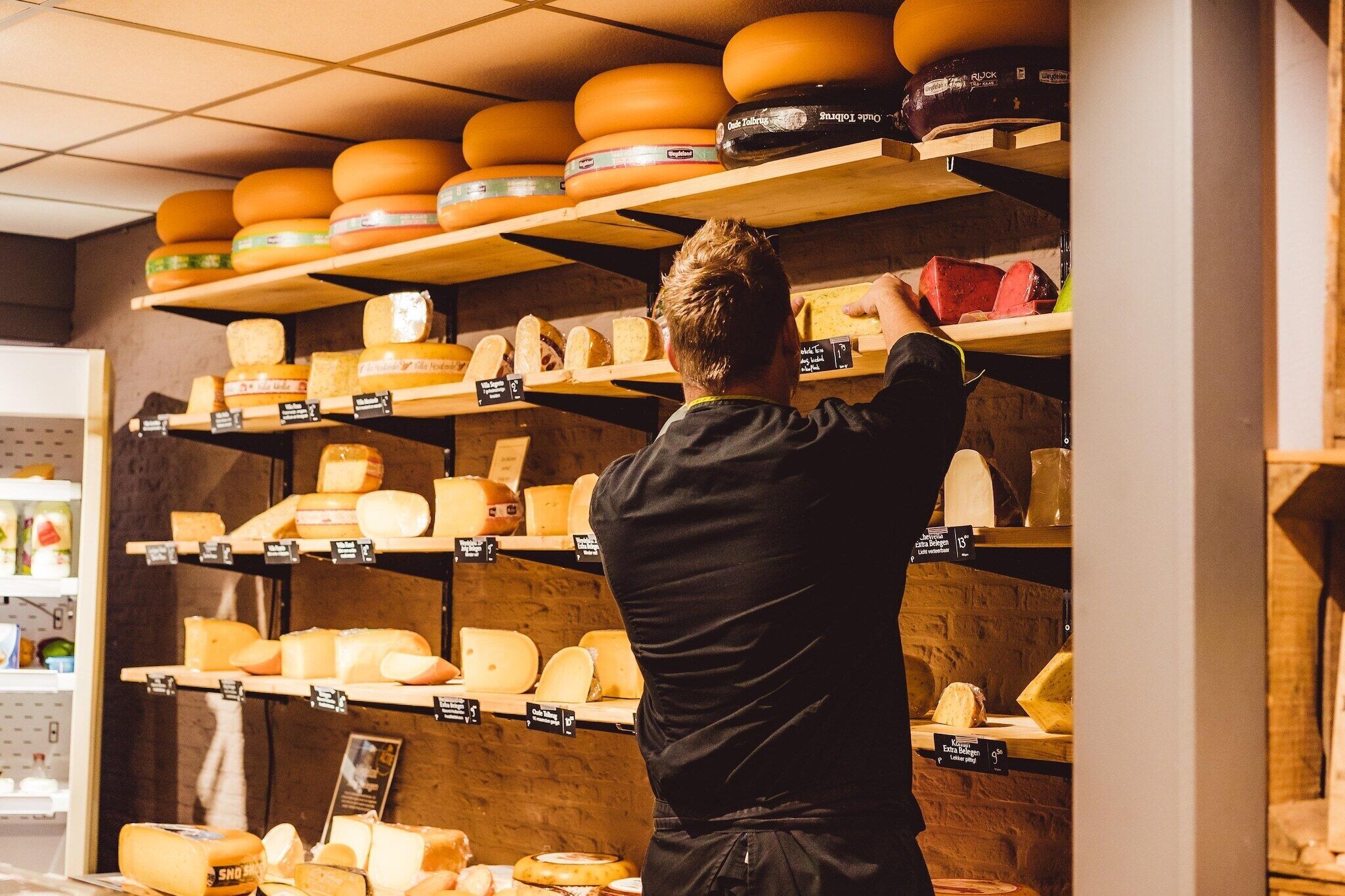 Une fromagerie avec des étagères remplies de différents fromages, et une personne qui tend la main pour en prendre un.