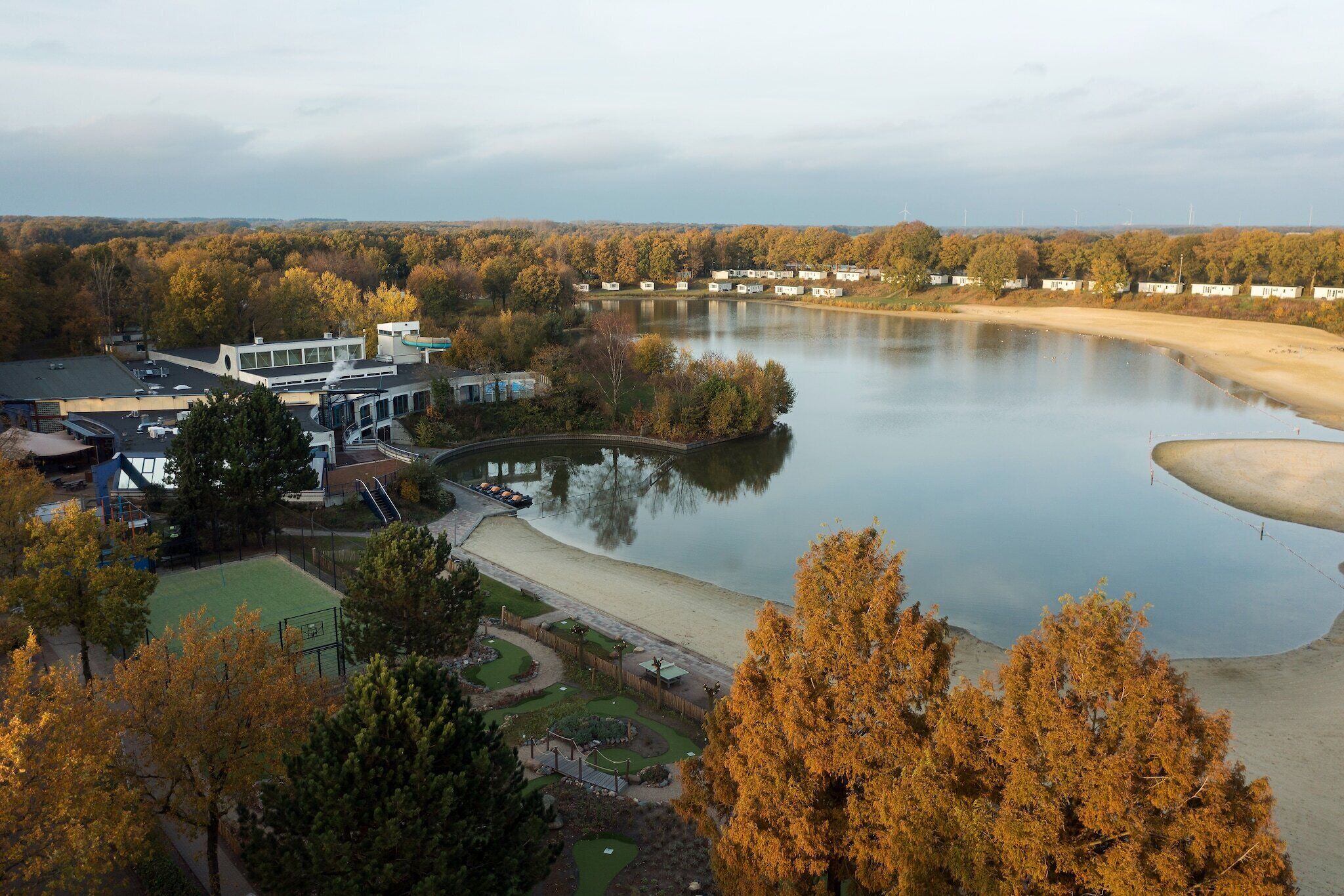 A lakeside view with a modern building, a sandy beach, and autumn-colored trees.