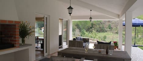 A covered patio area with a wooden table and benches, a brick fireplace, and a view of the surrounding landscape.