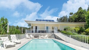Una piscina con sillas reclinables blancas, una escalera de piedra que lleva a un edificio con paneles solares y una valla blanca.