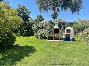A backyard with two outdoor pizza ovens, a wooden bench, and lush greenery.