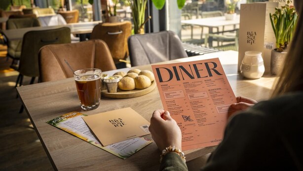 A person holding a menu at a restaurant table with a glass of beer, a plate of bread rolls, and a menu stand. - Bungalow in Bospark 't Wolfsven near Nature (Mierlo)