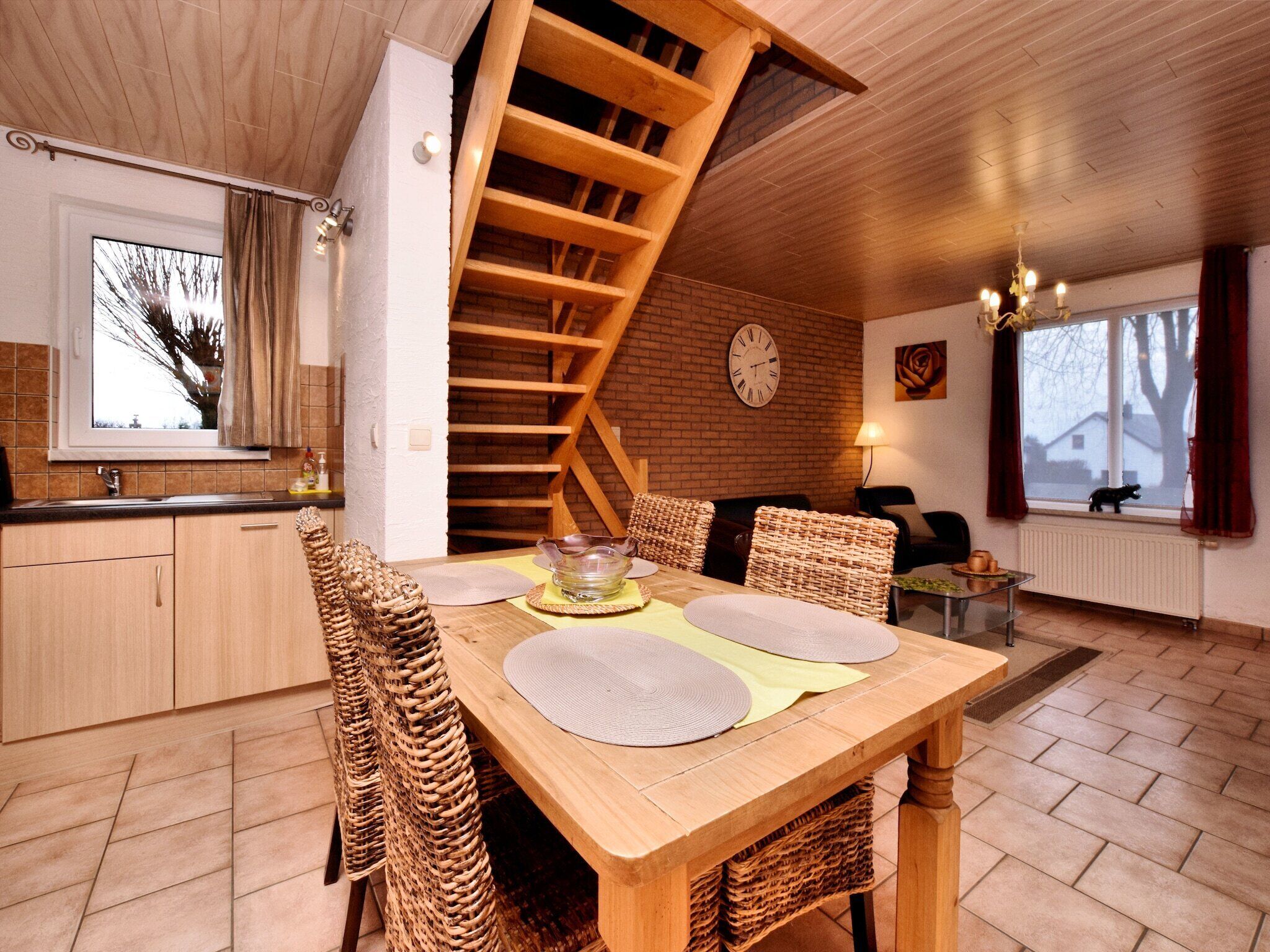 A dining area with a wooden table, wicker chairs, a brick wall, a clock, and a window with a view.