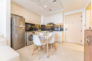 A modern kitchen with stainless steel appliances, a dining table with white chairs, and a washing machine. - Community View 1BR with Balcony Near Downtown Dubai, Binghatti Avenue, Al Jaddaf (Dubai)