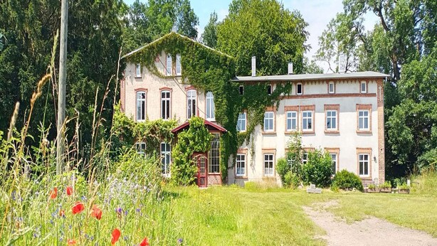 Une grande maison à deux étages, dotée d’un jardin à l’avant et entourée d’arbres.