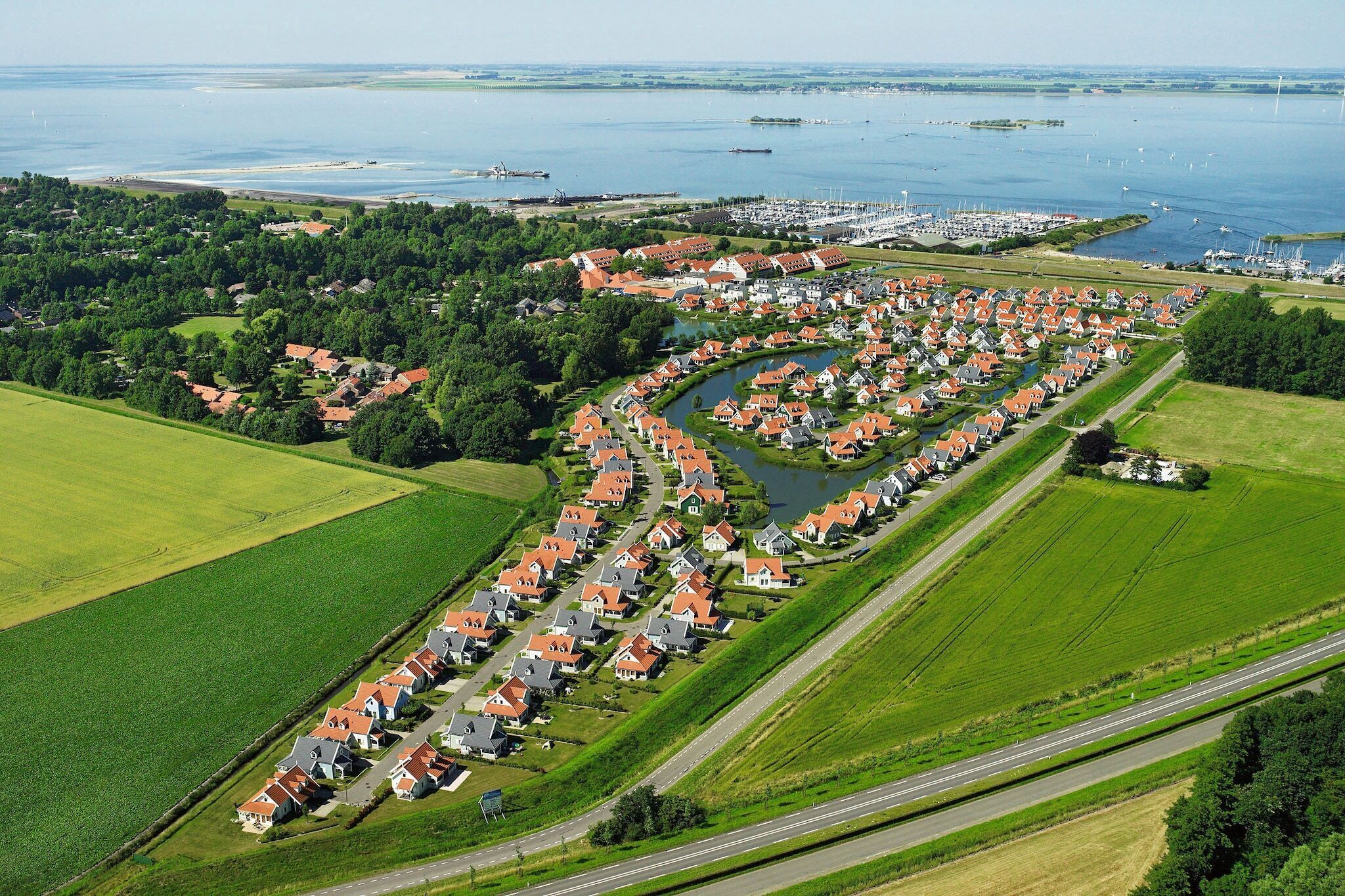 Vue aérienne d’un quartier résidentiel où les maisons sont alignées le long d’une rivière, avec un port de plaisance et des bâtiments industriels en arrière-plan.