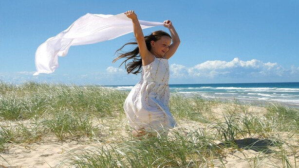 A person in a white dress with a flowing garment, standing on a sandy beach with grass and ocean in the background.