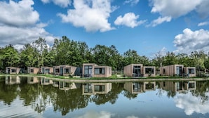 Uma fileira de cabanas modernas de madeira à beira de um lago tranquilo, rodeada por vegetação exuberante e um céu azul límpido.