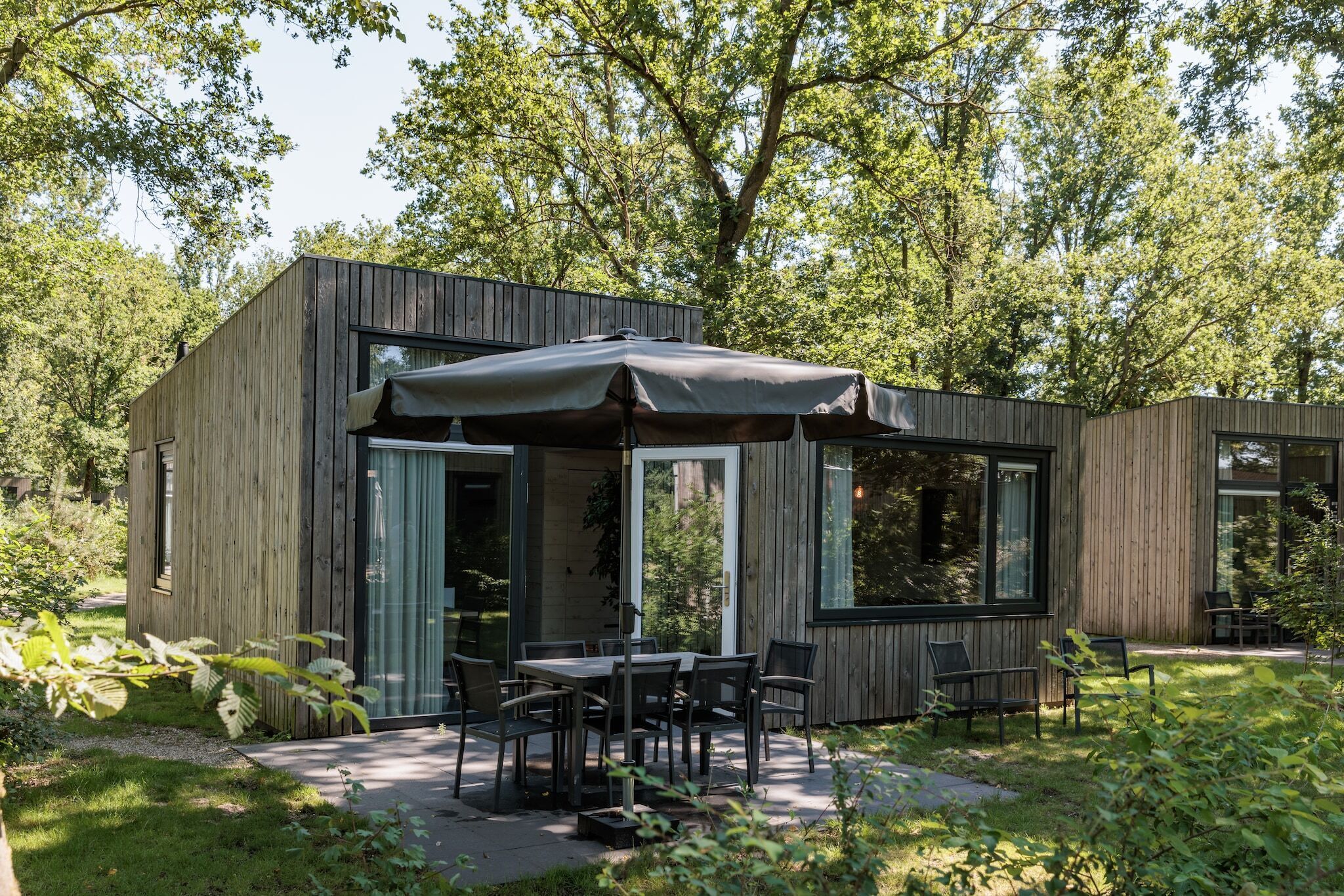 A modern wooden cabin with a patio area, including a table and chairs, surrounded by lush greenery.