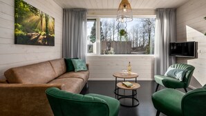 Sala de estar moderna com sofá, duas poltronas verdes, uma mesa redonda pequena e uma grande janela com vista para árvores e um edifício.